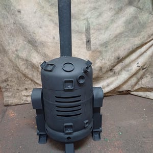 May include: A black metal fire pit shaped like a robot. The fire pit has a tall chimney and a round body with a grill-like opening. The robot has two legs and two arms.