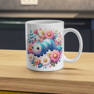 May include: A white ceramic mug with a colorful cartoon caterpillar surrounded by flowers. The caterpillar is blue, pink, and white with a cute face. The flowers are pink, white, and yellow.