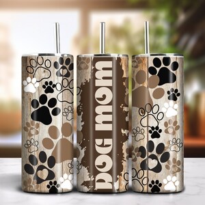 May include: Three stainless steel tumblers with paw prints and the text "DOG MOM" on a wood grain background. The paw prints are in black, white, and brown. Each tumbler has a silver straw.