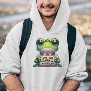 Puede incluir: Una sudadera con capucha blanca con una rana de dibujos animados verde que lleva corbata y sostiene un cartel que dice "No necesito suerte, tengo ranas".