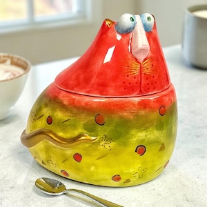 May include: A whimsical ceramic cat-shaped container with a lid. The cat's head is red, and the body is yellow and green with red and gold accents. A small spoon rests in front of the container. The container is likely used for storing sugar or other small items.