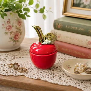May include: A glossy red ceramic apple-shaped trinket box with a metallic gold stem and leaf lid. The apple is positioned on a white lace doily, accompanied by two gold rings and a small cream-colored dish containing a pearl bracelet and a ring.