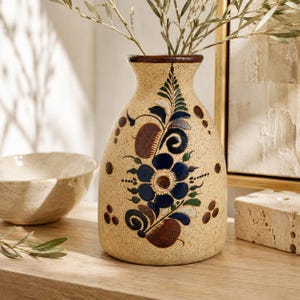 May include: A beige ceramic vase with a speckled texture and a dark brown rim. The front features a hand-painted floral design in shades of dark blue, brown, and green, with swirling leaf patterns and circular accents surrounding the central flower.