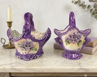 Könnte beinhalten: Zwei Keramikkörbe mit geschwungenen Henkeln, glänzend violetter Oberfläche mit weißen Punkten und handgemalten violetten Blumenclustern auf einem cremefarbenen Mittelteil. Die Ränder sind gewellt und mit erhabenen Verzierungen versehen.
