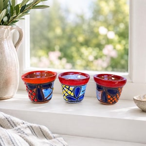 Op de afbeelding: Drie kleine, handbeschilderde keramische bekers met felrode randen. Elke beker heeft een uniek patroon van blauwe, gele en oranje geometrische vormen en stippen op een donkerblauwe achtergrond, opgesteld in een rij op een witte vensterbank.