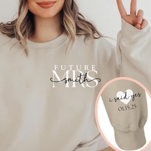 May include: Beige sweatshirt with the text "FUTURE MRS Smith" in a script font. The sleeve cuff features a heart and the words "i said yes" with the date 01.15.25. The person in the image is wearing a beige beanie.