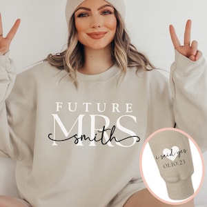 May include: A beige sweatshirt with the text "FUTURE MRS smith" in white lettering. The sleeve has a small white heart with the text "i said yes 01.10.23" in black lettering.