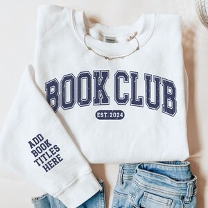 May include: White sweatshirt with the text "BOOK CLUB EST. 2024" in navy blue. The left sleeve has the text "ADD BOOK TITLES HERE" in navy blue.