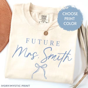 May include: A beige t-shirt with the text "FUTURE Mrs. Smith" in blue script with a blue bow underneath. The text "IVORY/MYSTIC PRINT" is at the bottom of the shirt.