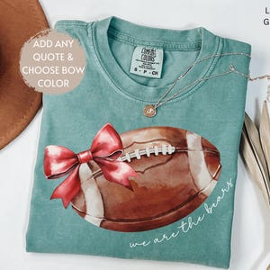 May include: A light green t-shirt featuring a watercolour illustration of a football with a red bow. The text "we are the bears" is printed on the shirt.