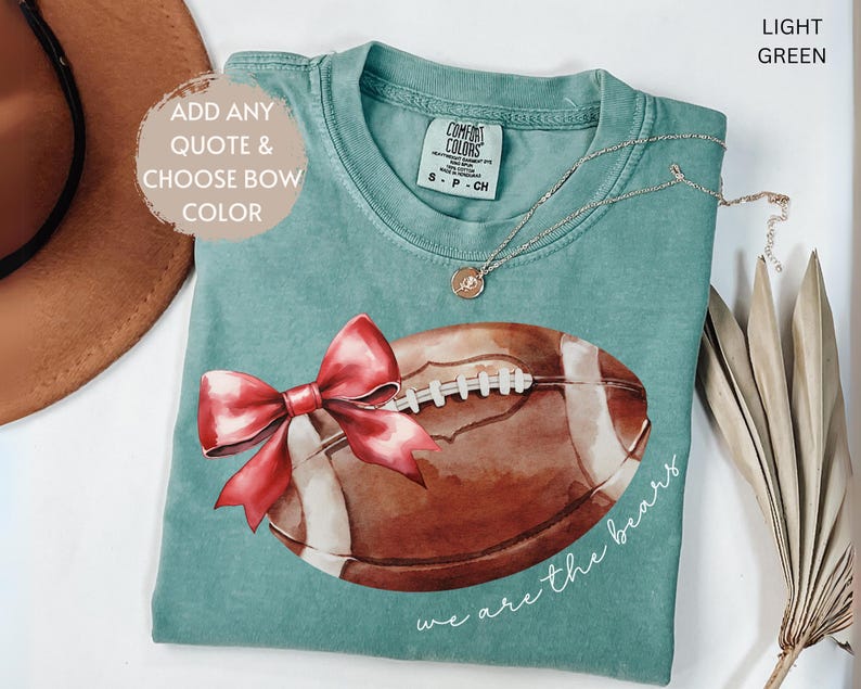 May include: A light green t-shirt featuring a watercolour illustration of a football with a red bow. The text "we are the bears" is printed on the shirt.