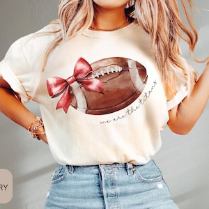 May include: Ivory t-shirt featuring a brown football and a red bow. The text "we are the titans" is printed on the shirt.
