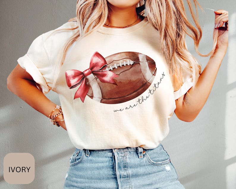 May include: Ivory t-shirt featuring a brown football and a red bow. The text "we are the titans" is printed on the shirt.