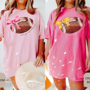 May include: Two pink t-shirts with a white football graphic and the text "Let's go Spartans". The shirt on the left has a pink bow on the football and the shirt on the right has a yellow bow on the football.