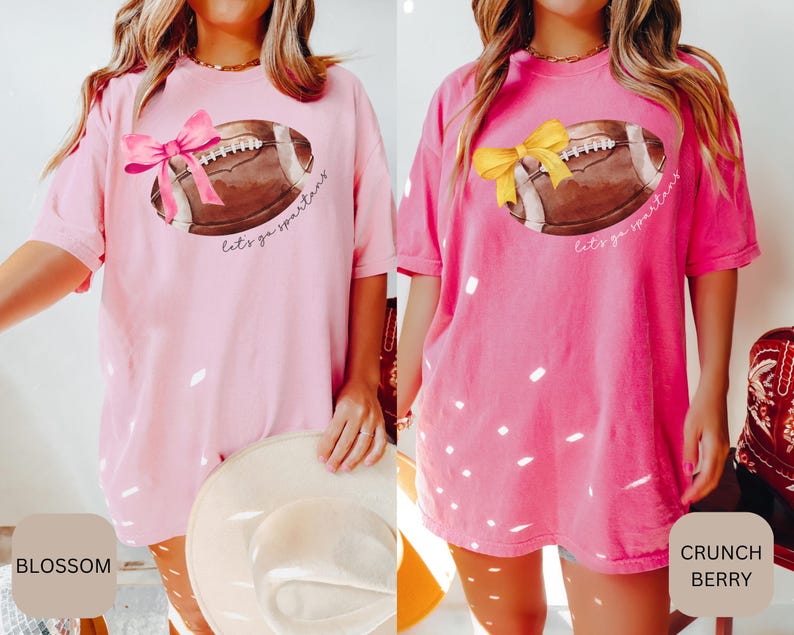 May include: Two pink t-shirts with a white football graphic and the text "Let's go Spartans". The shirt on the left has a pink bow on the football and the shirt on the right has a yellow bow on the football.
