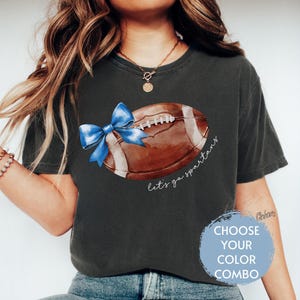 May include: A dark grey t-shirt featuring a watercolour illustration of a football with a blue bow on it. The text "Let's go Spartans" is written below the football.