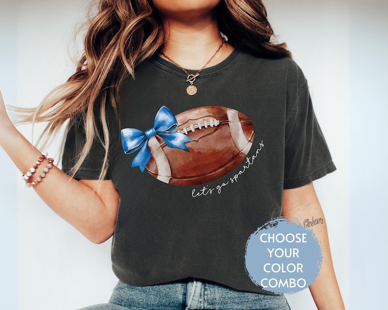 May include: A dark grey t-shirt featuring a watercolour illustration of a football with a blue bow on it. The text "Let's go Spartans" is written below the football.