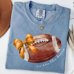 May include: A blue jean t-shirt featuring a watercolour illustration of a football and a yellow bow. The text "we are the bears" is printed on the shirt.