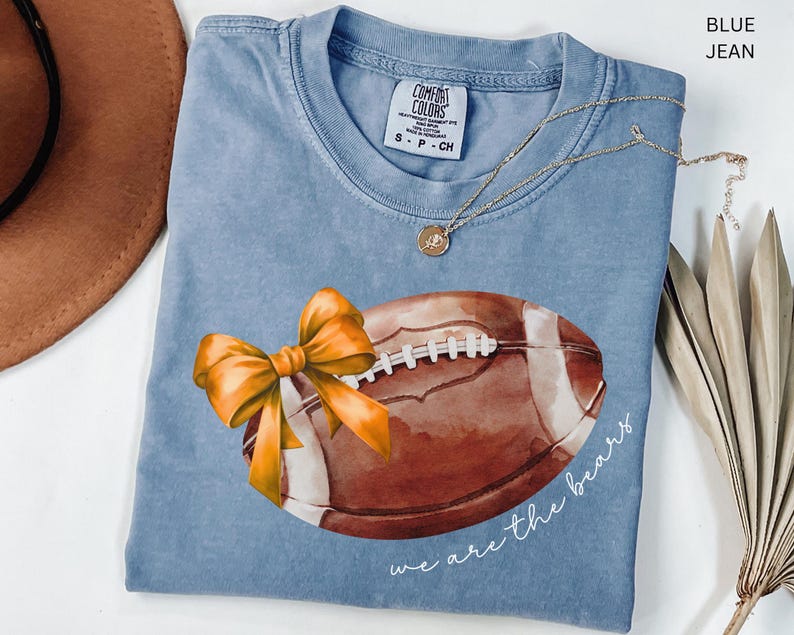May include: A blue jean t-shirt featuring a watercolour illustration of a football and a yellow bow. The text "we are the bears" is printed on the shirt.