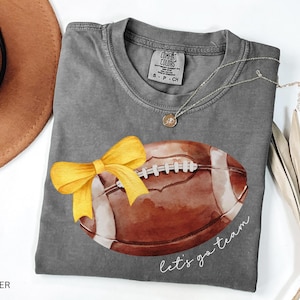 May include: Grey t-shirt with a watercolour illustration of a football with a yellow bow. The text "Let's go team" is printed on the shirt.