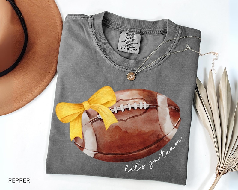 May include: Grey t-shirt with a watercolour illustration of a football with a yellow bow. The text "Let's go team" is printed on the shirt.