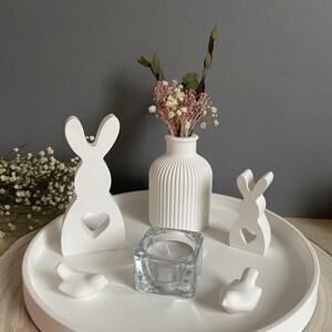 Könnte beinhalten: Weiße Keramik-Schale mit zwei weißen Hasenfiguren, einer weißen Vase mit Trockenblumen, einem durchsichtigen Glas-Kerzenhalter und zwei weißen Vogelfiguren.