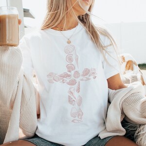May include: A white t-shirt with a pink seashell cross design. The shirt is being worn by a person sitting on a white fluffy chair.