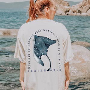 May include: Ivory-colored t-shirt with a blue and white stingray design on the back. The text around the stingray reads "WHEN YOU GO THROUGH DEEP WATERS, I WILL BE WITH YOU." The shirt is worn outdoors near a body of water.