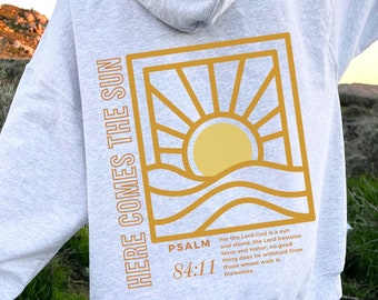 Könnte beinhalten: Grauer Hoodie mit einer Grafik einer Sonne, die über Wellen aufgeht, in einem goldenen Rahmen. Der Text "HERE COMES THE SUN" ist vertikal auf der linken Seite des Rahmens gedruckt. Der Text "PSALM 84:11" ist unter dem Rahmen gedruckt. Der Text "Denn der Herr Gott ist Sonne und Schild; der Herr gibt Gnade und Ehre; er wird denen, die in Rechtschaffenheit wandeln, nichts Gutes versagen." ist unter dem Text "PSALM 84:11" gedruckt.