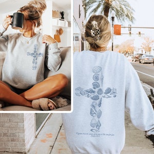 May include: A light gray sweatshirt featuring a cross design made of seashells. The sweatshirt has long sleeves and a crew neck. The text "Tighter than the waves of the sea is His love for you. Psalm 93:4" is printed below the cross.