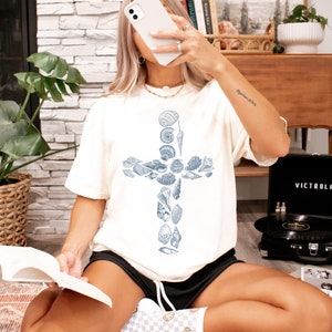 May include: A white t-shirt with a blue seashell cross design. The shirt is being worn by a person sitting on a rug with a book in their lap. The text "ivory" is visible in the upper right corner of the image.