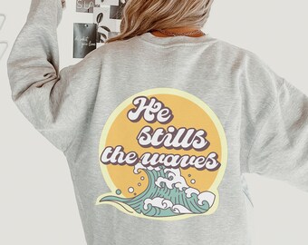 Könnte beinhalten: Ein graues Sweatshirt mit einem gelben Kreis und einer blauen Wellengrafik. Der Text "He stills the waves" ist in Braun auf dem Kreis gedruckt.