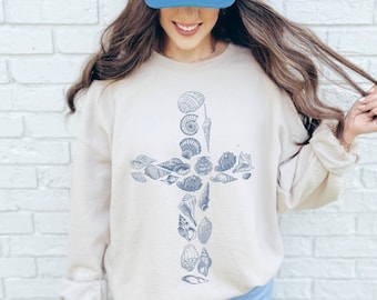 May include: A beige sweatshirt with a blue seashell cross design. The model is wearing a blue trucker hat.