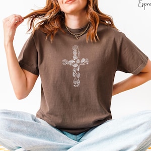 May include: A brown t-shirt with a white cross made of seashells. The cross is centered on the shirt.