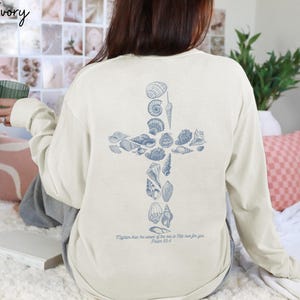 May include: Ivory long-sleeve shirt with a blue seashell cross design on the back. The shirt has the text "More than the sands of the sea is His love for you. Psalm 93:4" printed below the cross.