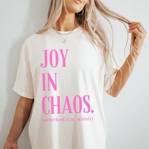 May include: White t-shirt with the text "JOY IN CHAOS. motherhood is my ministry" printed in pink.