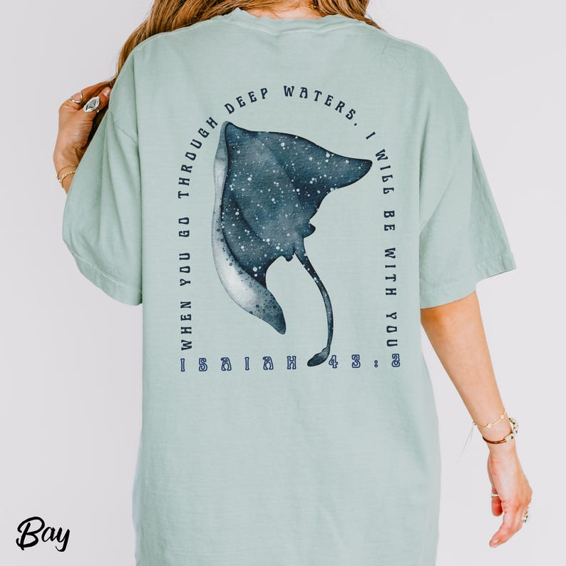 May include: A light green t-shirt with a watercolor illustration of a manta ray. The design includes the text "WHEN YOU GO THROUGH DEEP WATERS. I WILL BE WITH YOU" and "ISAIAH 43:2". The word "Bay" is printed at the bottom.