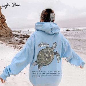 May include: Light blue hoodie with a sea turtle graphic on the back. The hoodie has a drawstring hood and a kangaroo pocket. The text on the back reads "THE FISH IN THE SEA, ALL THAT SWIM THE PATHS OF THE SEAS. LORD, OUR LORD, HOW MAJESTIC IS YOUR NAME IN ALL THE EARTH! PSALM 8:8-9"