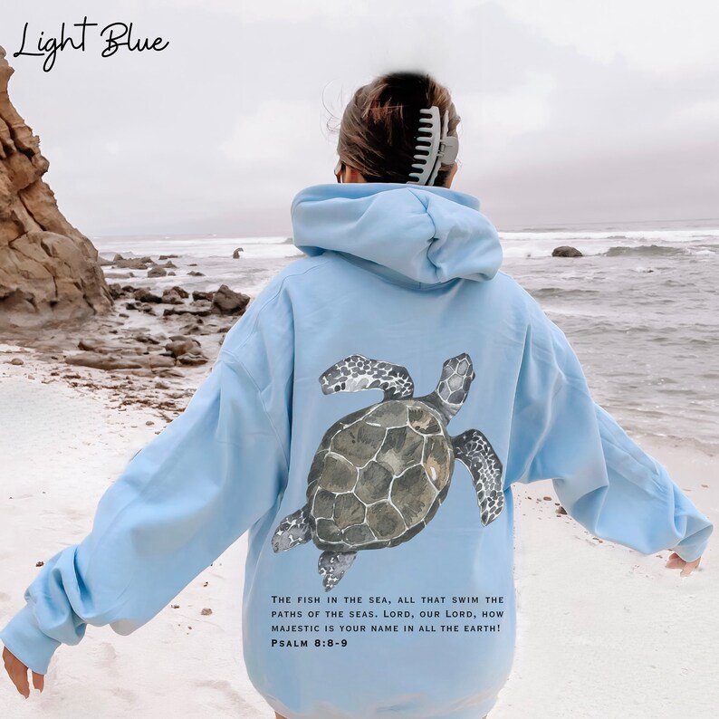 May include: Light blue hoodie with a sea turtle graphic on the back. The hoodie has a drawstring hood and a kangaroo pocket. The text on the back reads "THE FISH IN THE SEA, ALL THAT SWIM THE PATHS OF THE SEAS. LORD, OUR LORD, HOW MAJESTIC IS YOUR NAME IN ALL THE EARTH! PSALM 8:8-9"