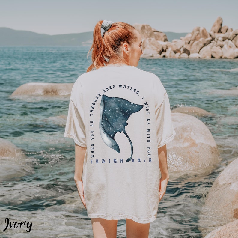 May include: Ivory-colored t-shirt with a blue and white stingray design on the back. The text around the stingray reads "WHEN YOU GO THROUGH DEEP WATERS, I WILL BE WITH YOU." The shirt is worn outdoors near a body of water.
