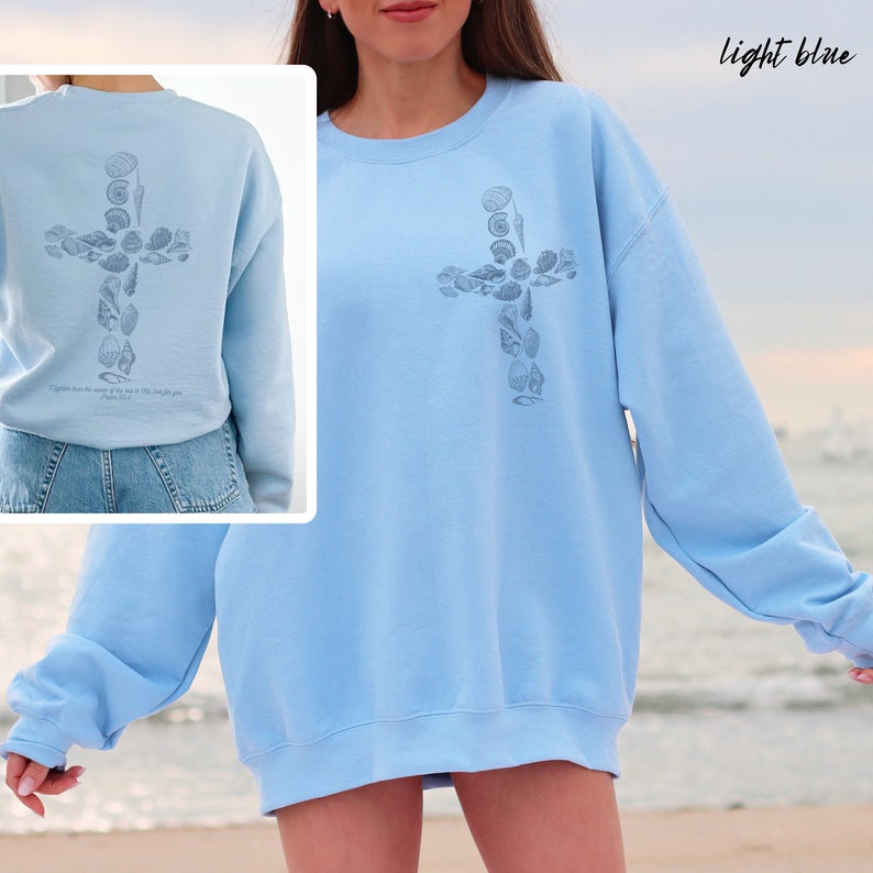 May include: Light blue sweatshirt featuring a cross design made of seashells. The back of the sweatshirt also has the cross design and the text "Psalm 91:1". The words "light blue" are written in cursive at the top right.