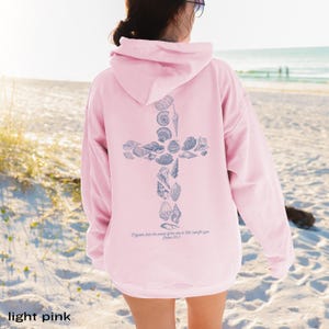 May include: Light pink hoodie with a cross design made of seashells. The back of the sweatshirt features the text "Mightier than the waves of the sea is His love for you, Psalm 93:4."