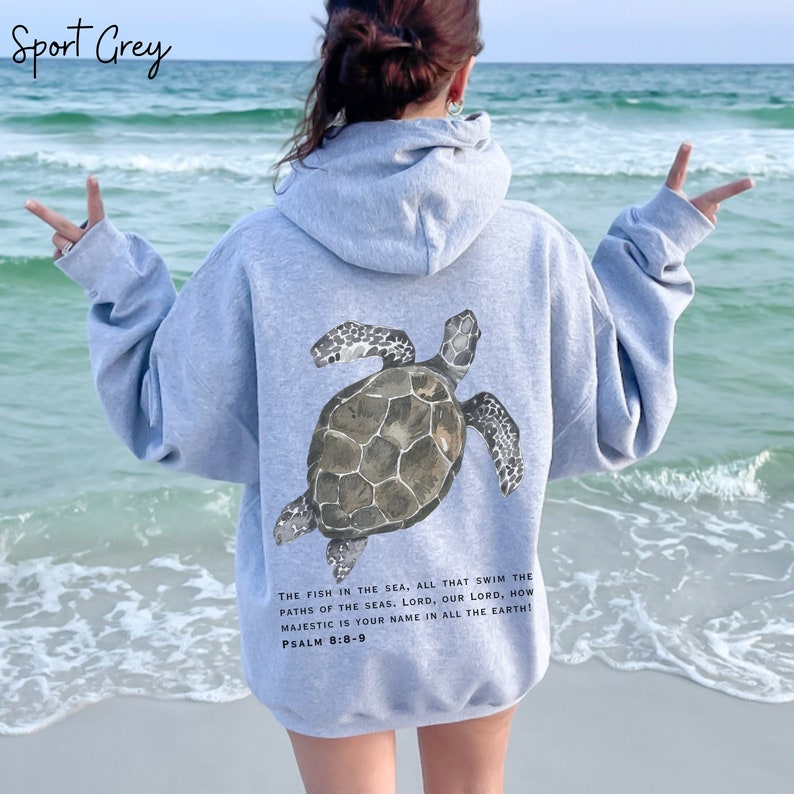 May include: A light gray hooded sweatshirt with a sea turtle graphic on the back. The turtle is brown and gray with a detailed shell. The text "THE FISH IN THE SEA, ALL THAT SWIM THE PATHS OF THE SEAS. LORD, OUR LORD, HOW MAJESTIC IS YOUR NAME IN ALL THE EARTH! PSALM 8:8-9" is printed below the turtle.