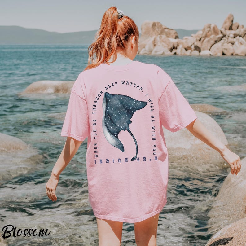 May include: Pink t-shirt with a large illustration of a manta ray and the text "When you go through deep waters, I will be with you." The manta ray is dark blue with white accents. The word "Blossom" is in the bottom left corner.
