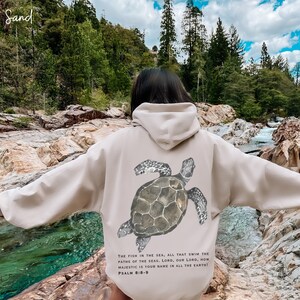 May include: A beige hoodie with a watercolor illustration of a sea turtle on the back. The hoodie has a drawstring hood and a kangaroo pocket. The text "THE FISH IN THE SEA, ALL THAT SWIM THE PATHS OF THE SEAS. LORD, OUR LORD, HOW MAJESTIC IS YOUR NAME IN ALL THE EARTH! PSALM 8:8-9" is printed below the turtle.