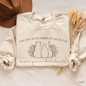 May include: A beige sweatshirt with a black graphic of three vases with floral accents. The text "Like clay in the hands of the potter so are you in God's hands. Jeremiah 18:6" is printed on the sweatshirt.