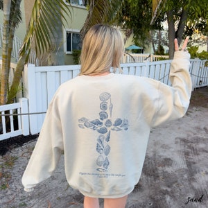 May include: Sand-colored sweatshirt with a blue seashell cross design on the back. The text "Mightier than the waves of the sea is His love for you. Psalm 93:4" is printed below the cross. The sweatshirt is a relaxed fit.