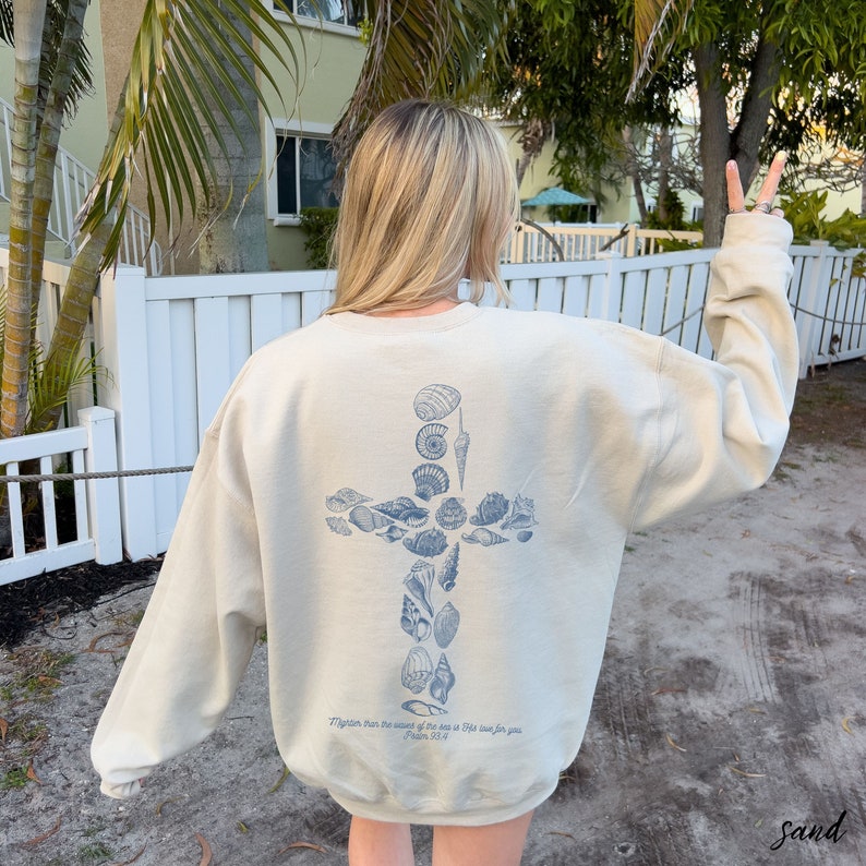 May include: Sand-colored sweatshirt with a blue seashell cross design on the back. The text "Mightier than the waves of the sea is His love for you. Psalm 93:4" is printed below the cross. The sweatshirt is a relaxed fit.