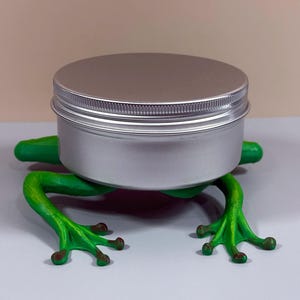 May include: A silver metal jar with a ribbed lid sits atop a bright green frog-shaped holder. The frog has detailed feet and a textured appearance. The background is a soft beige and white.