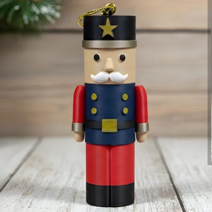 May include: A wooden nutcracker figurine in a soldier's uniform. The figurine has a black hat with a gold star, a navy blue jacket, red trousers, and a white moustache. A smaller version of the nutcracker is also shown.